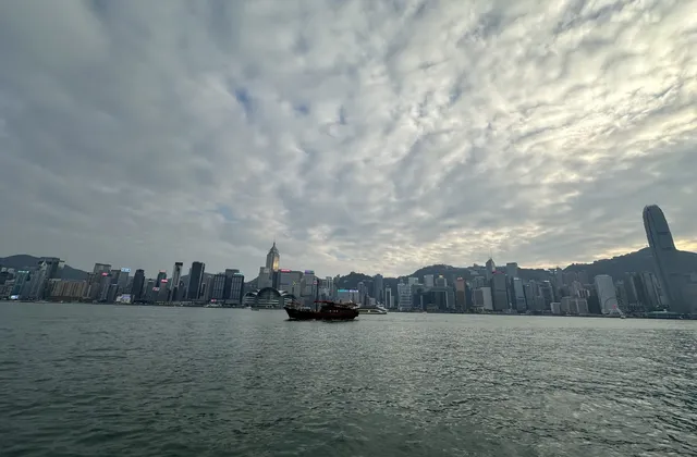Victoria Harbour and the Hong Kong Island skyline under a wide cloudy sky, with a junk on the water