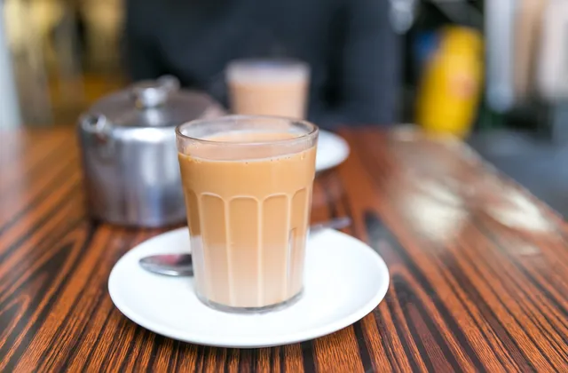 A glass of milk tea on a saucer beside a metal teapot