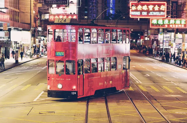 A red double-decker tram at night on a wide street lined with neon signs